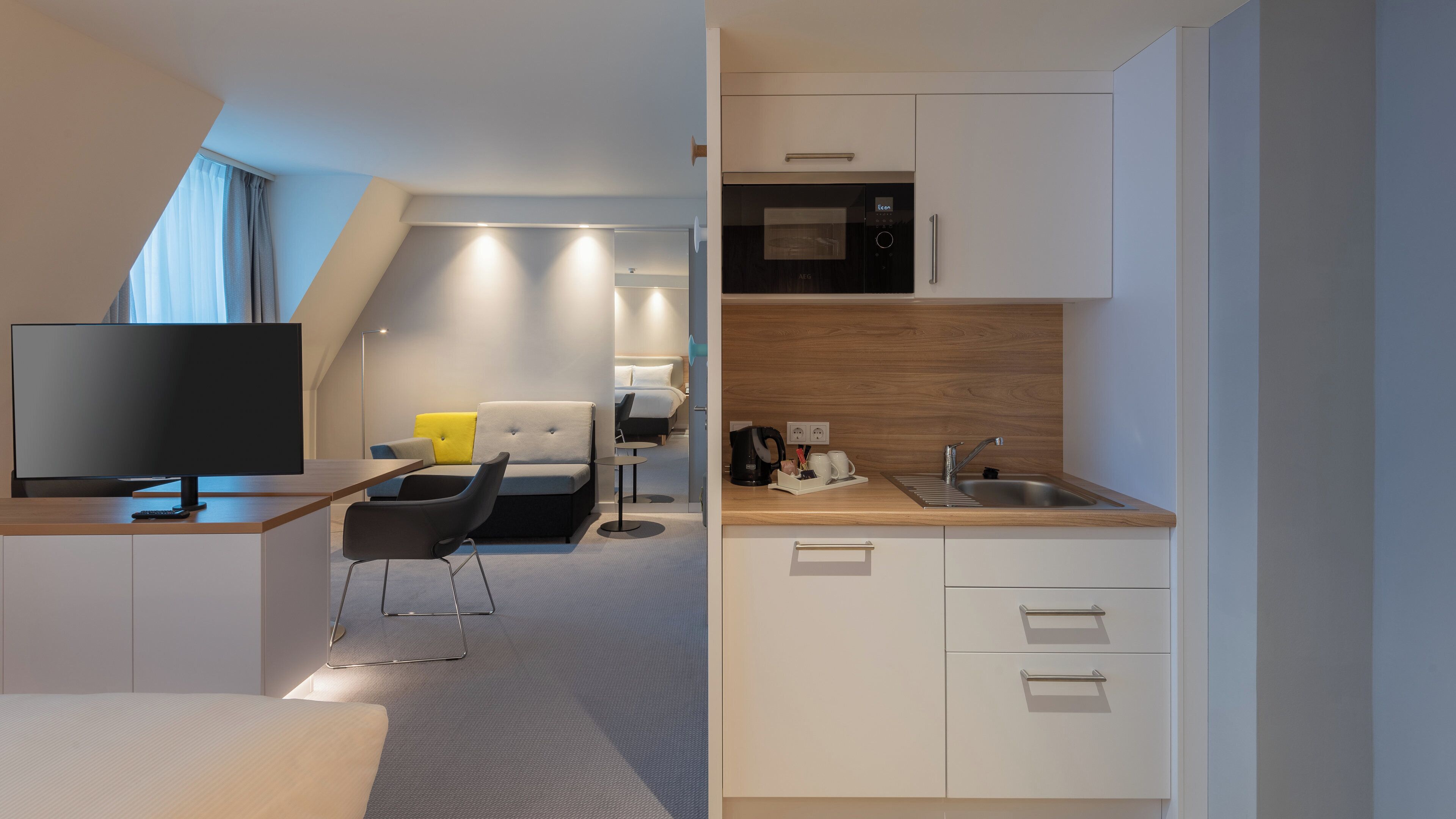 A modern hotel room with a flat-screen TV, a sofa, a small dining table, and a kitchenette with a microwave and sink.