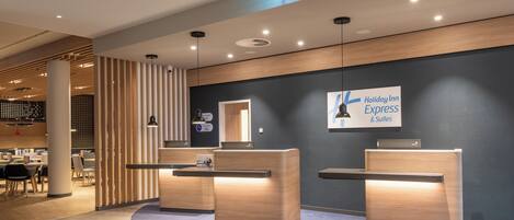 Una moderna zona de recepción de hotel con dos mostradores, un gran letrero de Holiday Inn Express & Suites, y un área de descanso con mesas y sillas.