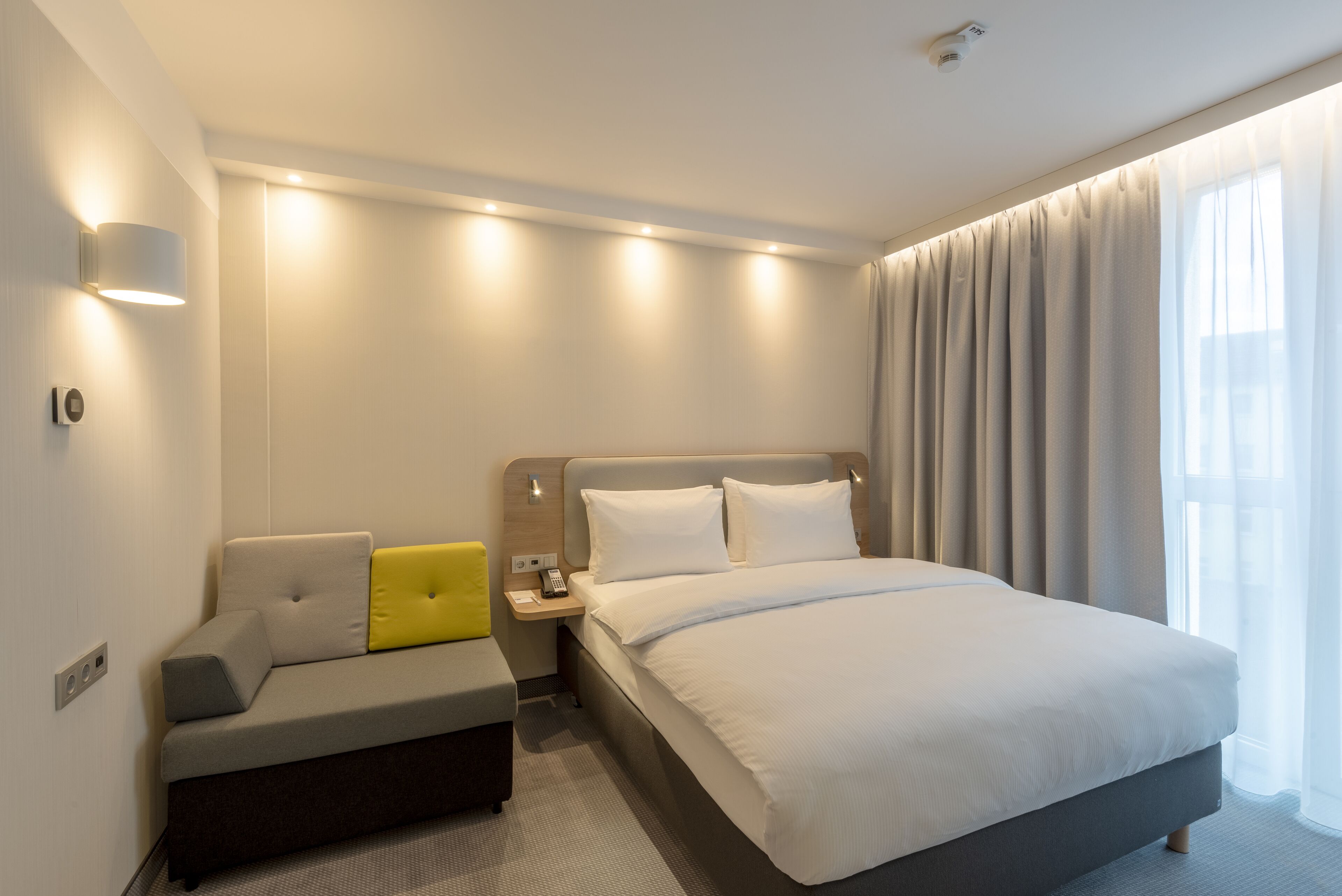 Una moderna camera d'hotel con un letto grande, un divano, un comodino con una lampada e una finestra con tende.