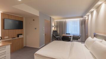 Ett hotellrum med en stor säng, ett skrivbord med en stol, en TV som är monterad på väggen och ett fönster med gardiner.