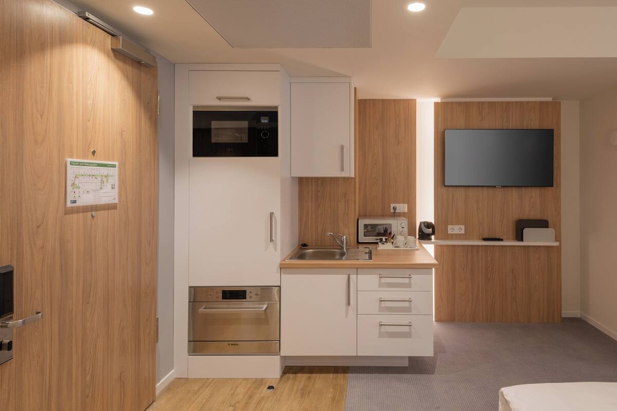 A modern kitchen with a Bosch oven, microwave, and sink. There is a flat-screen TV mounted on the wall and a small shelf with a kettle and cups.