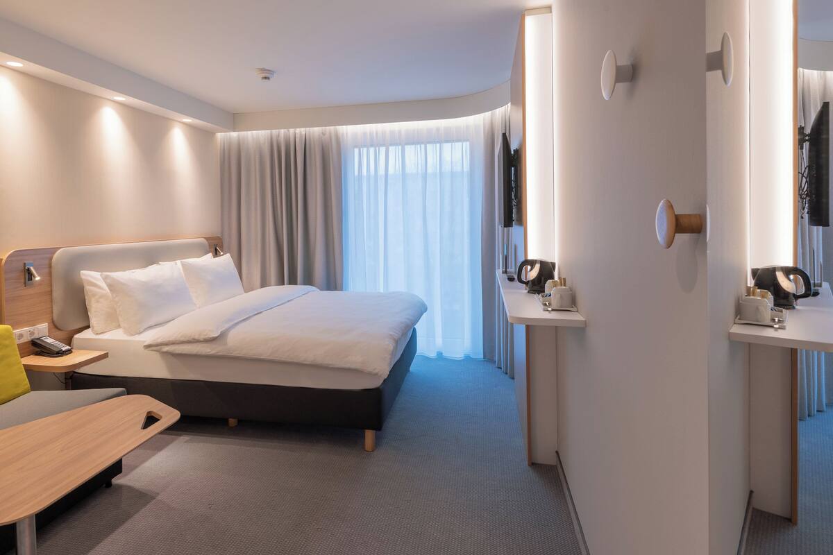 A modern hotel room with a neatly made bed, a small desk, a sofa, and a bathroom area visible through an open door.