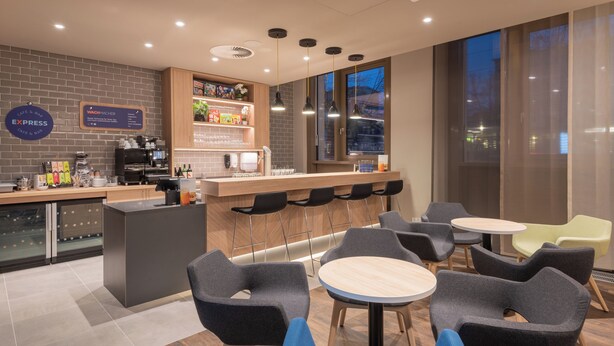 A modern cafe with a bar area, seating, and a view of the city at dusk. - Holiday Inn Express and Suites Potsdam by IHG (Potsdam)