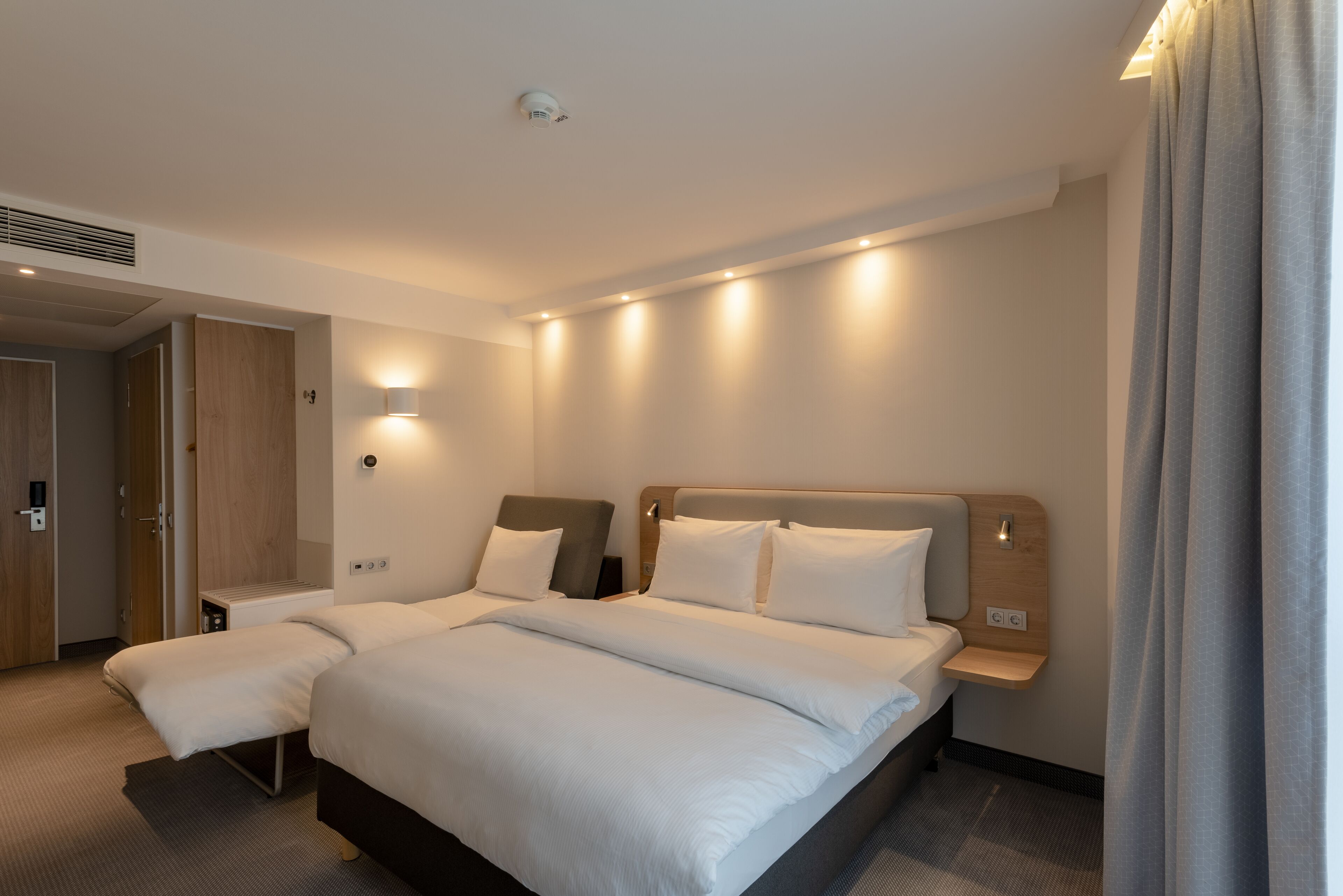A modern hotel room with a large bed, bedside tables, a built-in shelf, and a ceiling-mounted light fixture.