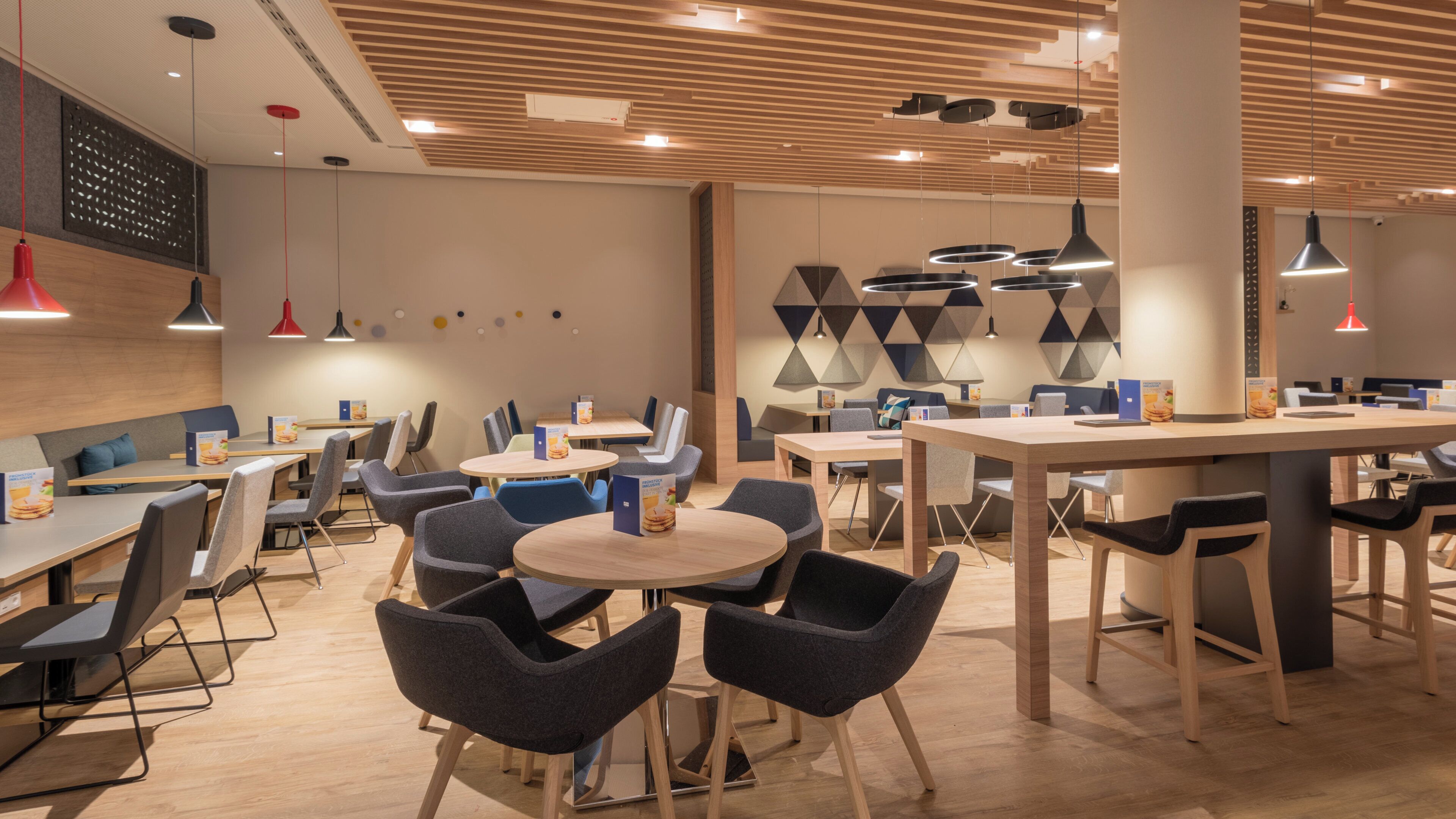 Un restaurant moderne avec des tables rondes et des chaises grises, agrémenté d’œuvres d’art géométriques sur les murs et de luminaires suspendus.