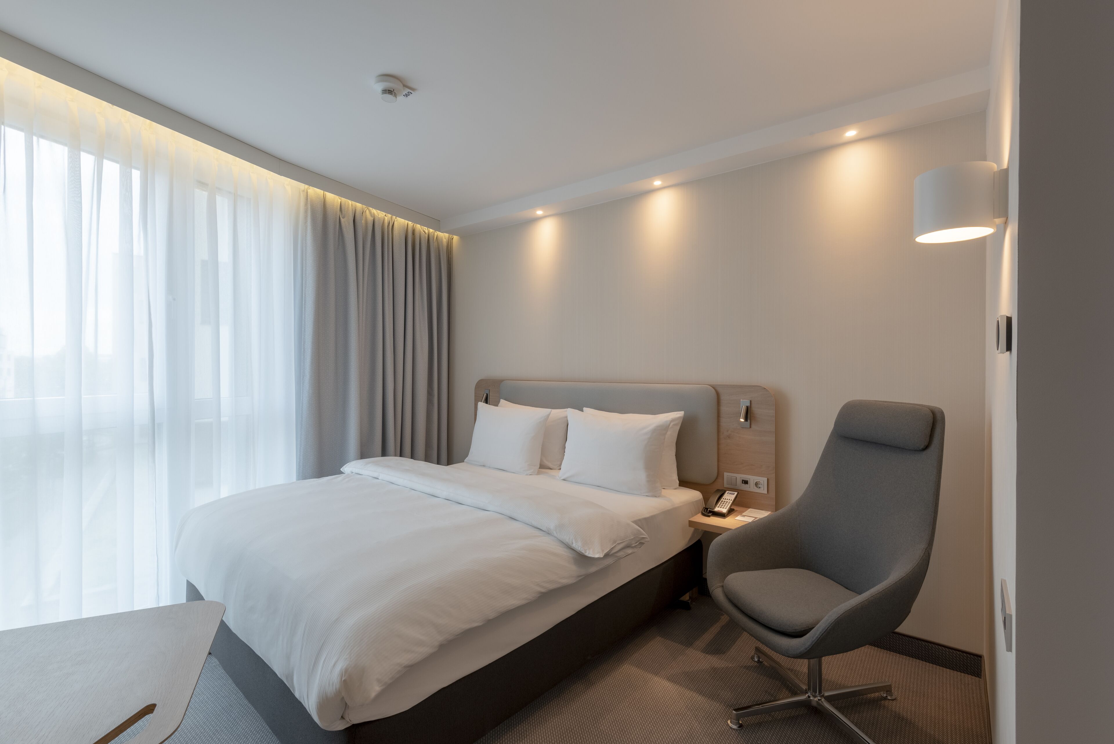 Una habitación de hotel moderna con una cama bien tendida, una mesita de noche con una lámpara y una silla.