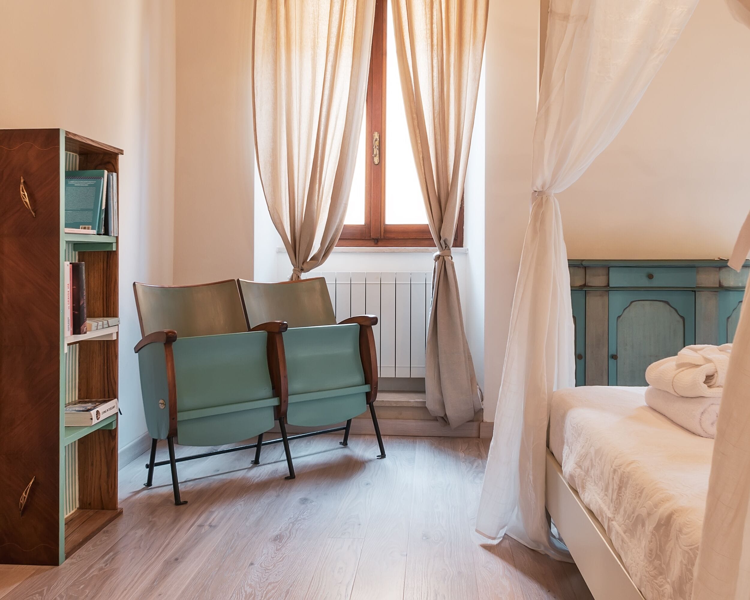 Ein Schlafzimmer mit einem Bett, einem Sessel, einem Bücherregal und einem Fenster mit Vorhängen.