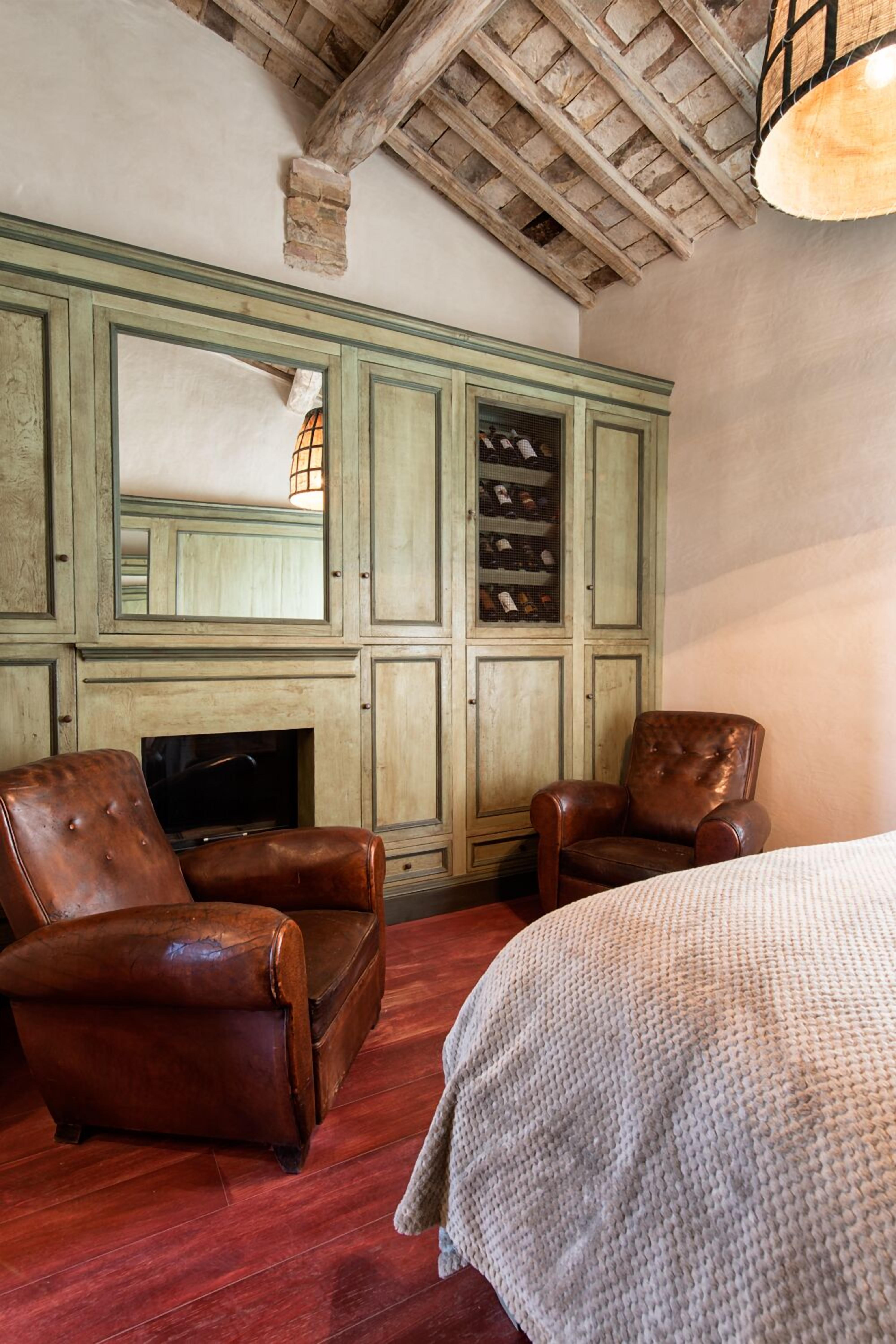 A room with a wooden ceiling, a green cabinet with wine bottles, leather chairs, and a bed with a white blanket.