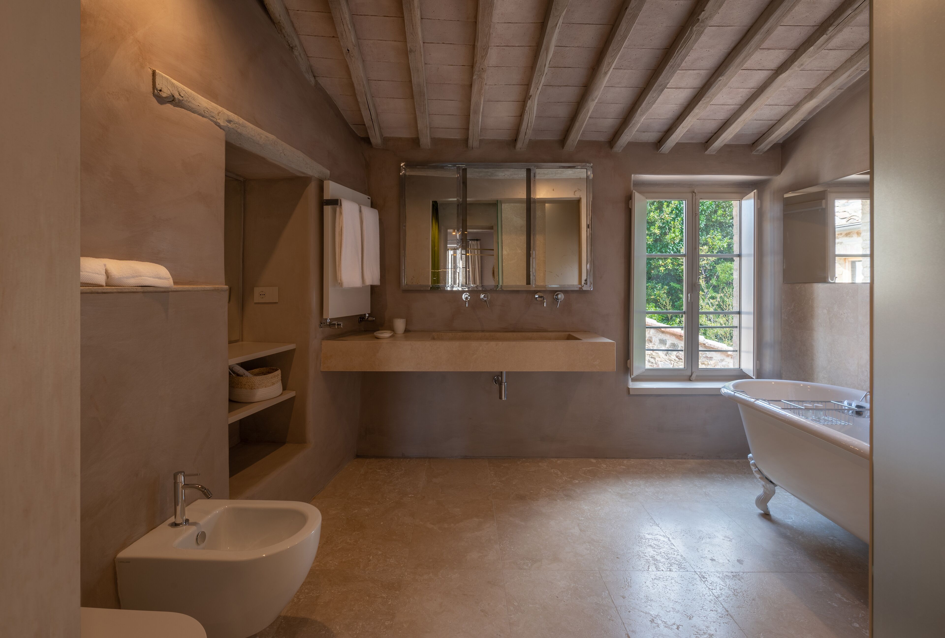 A bathroom with a freestanding bathtub, a sink with a mirror above it, and a bidet. There is a window providing natural light and a view of greenery outside.