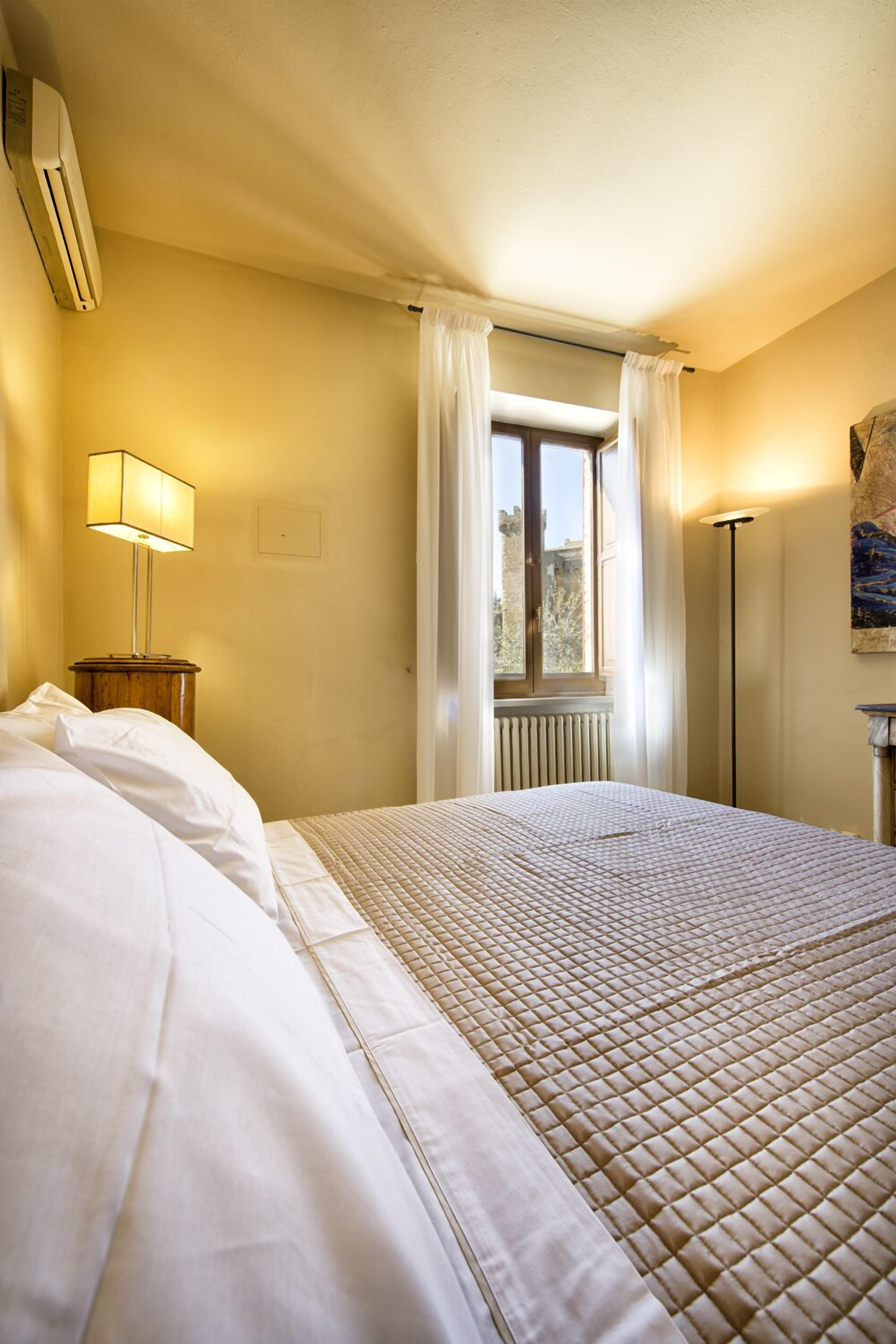 A hotel room with a bed, a lamp, a radiator, and a window with curtains.