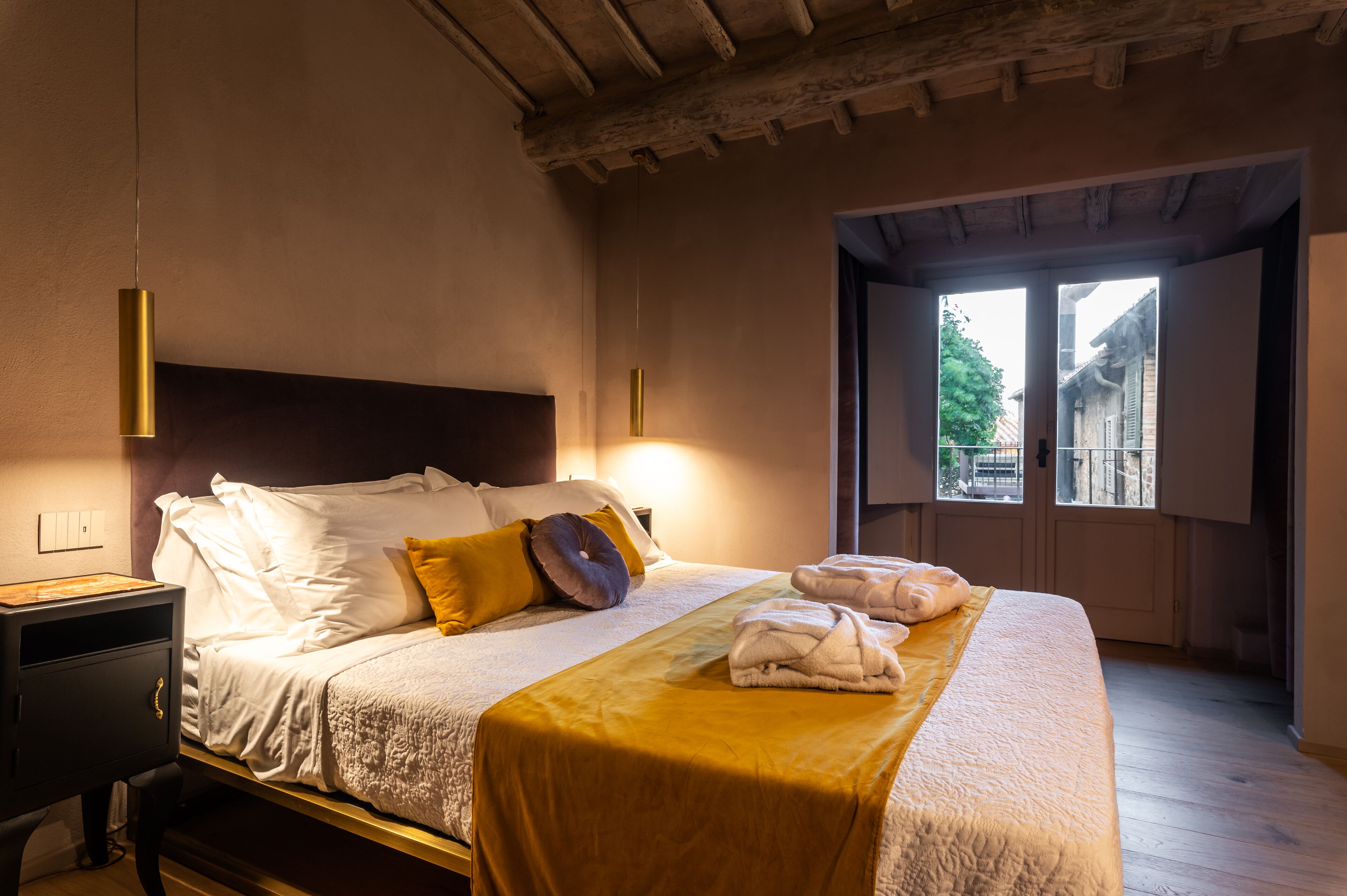 Un lit bien fait, avec du linge de lit blanc et un plaid doré, une table de chevet avec une lampe, et une vue sur l’extérieur par une porte.