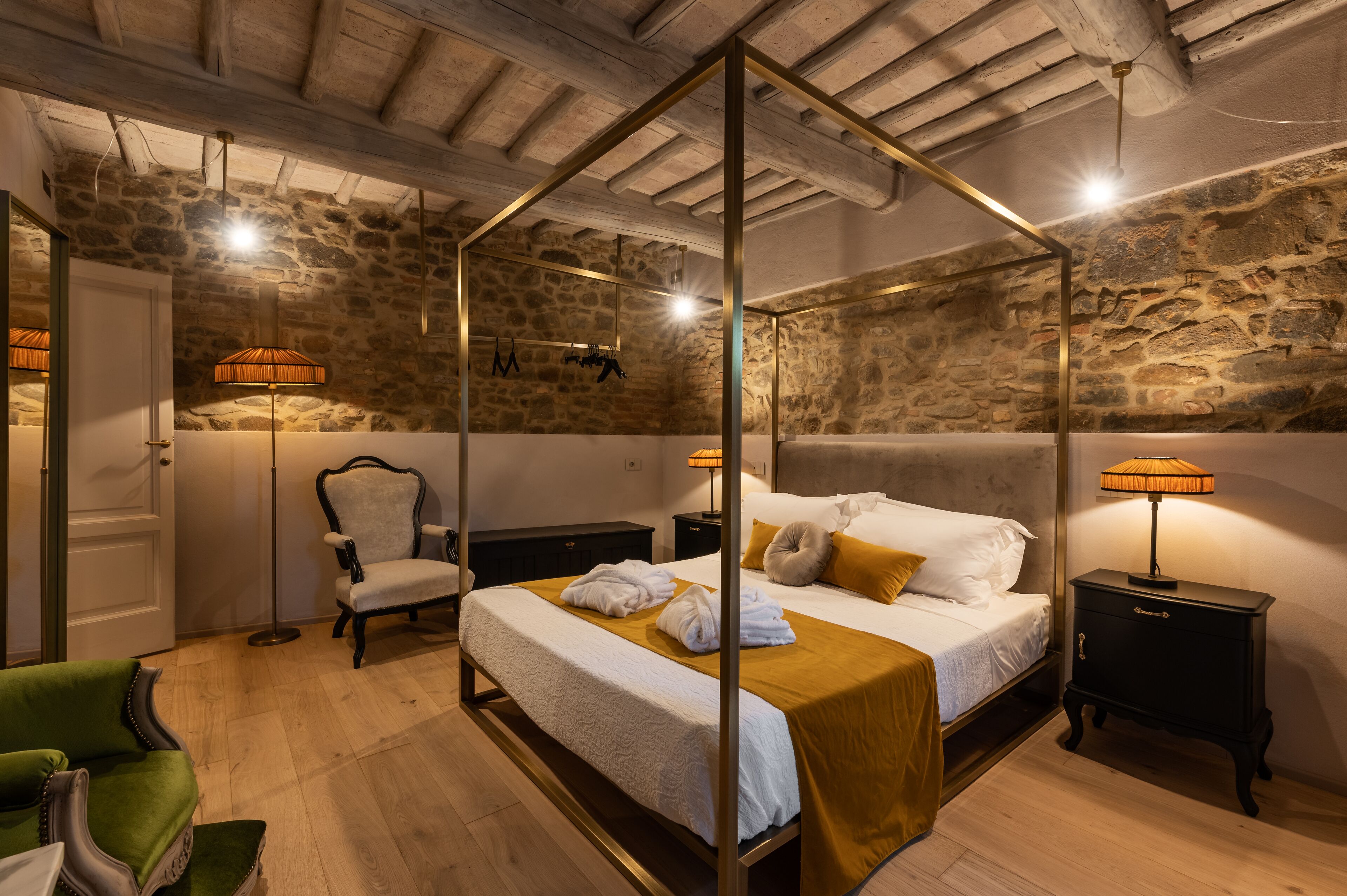 A four-poster bed with a white bedspread and gold throw, a green armchair, a nightstand with a lamp, and a stone wall.