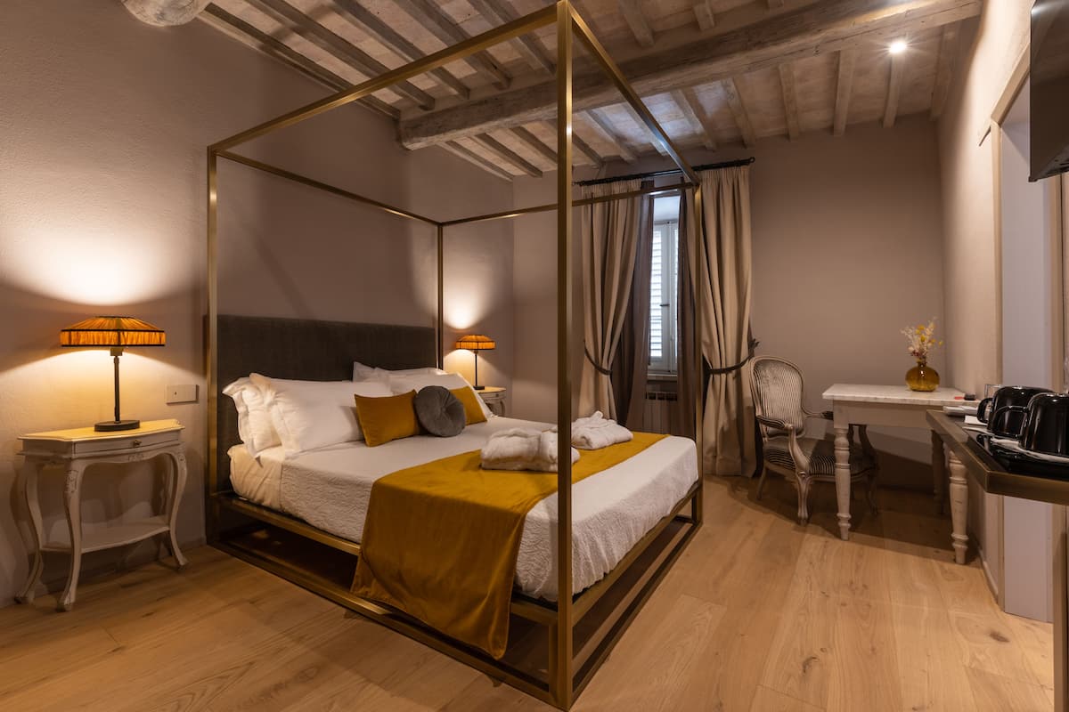 Una cama con dosel con ropa de cama blanca y color mostaza, una mesita de noche con una lámpara, una silla y una pequeña mesa con un florero y una tetera.