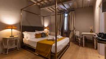 A four-poster bed with a white and mustard-colored bedding, a small bedside table with a lamp, a chair, and a small table with a vase and a kettle.