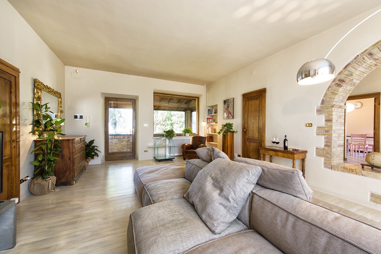 A spacious living room with a large sofa, wooden furniture, and a view of the outdoors through a glass door.