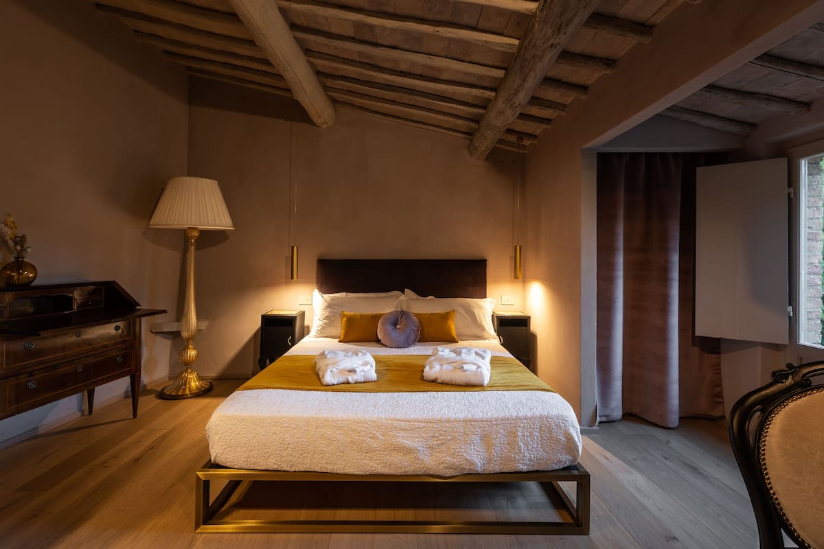 Habitación de hotel con techo de madera, cama con ropa de cama blanca, dos toallas, una mesita de noche, una lámpara y una ventana con cortinas.