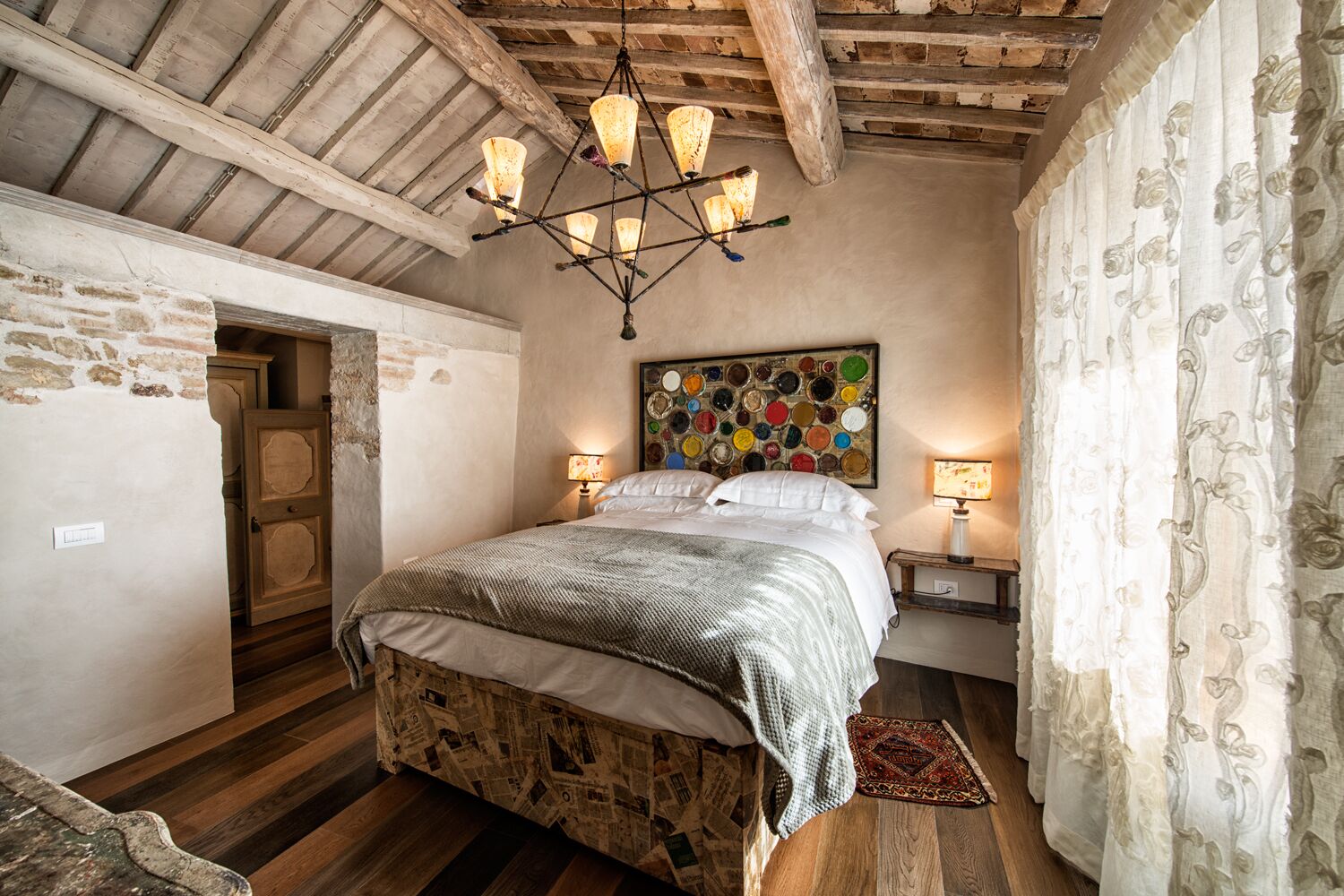 Quarto com teto de madeira, cama com colcha estampada, uma obra de arte colorida na parede e duas luminárias de cabeceira.
