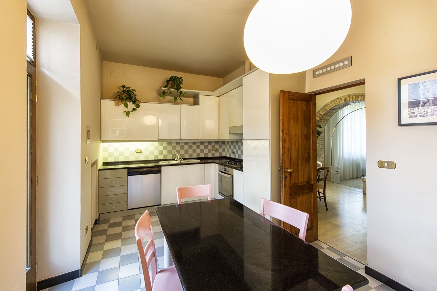 Une cuisine moderne avec des meubles blancs, un plan de travail noir et un dosseret à carreaux. On y trouve une table à manger avec des chaises roses et une grande fenêtre qui laisse entrer la lumière naturelle.