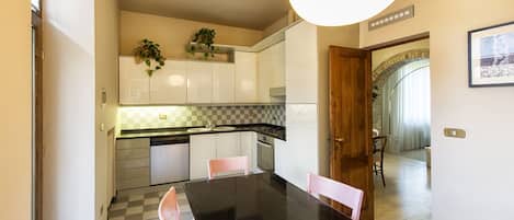 A modern kitchen with white cabinets, a black countertop, and a checkered backsplash. There is a dining table with pink chairs and a large window providing natural light.