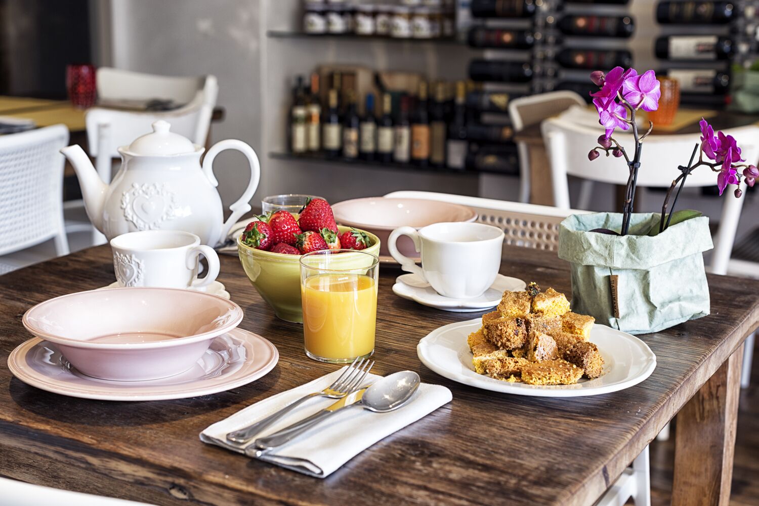Une table de petit-déjeuner avec une théière, des tasses, un bol de fraises, un verre de jus d’orange et une assiette de nourriture.