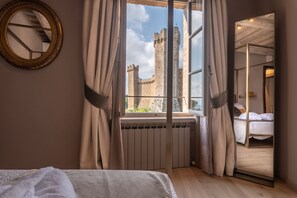 Enjoy a room with a large window offering a view of a historic castle, featuring a comfortable bed, a round mirror, and a radiator beneath the window.
