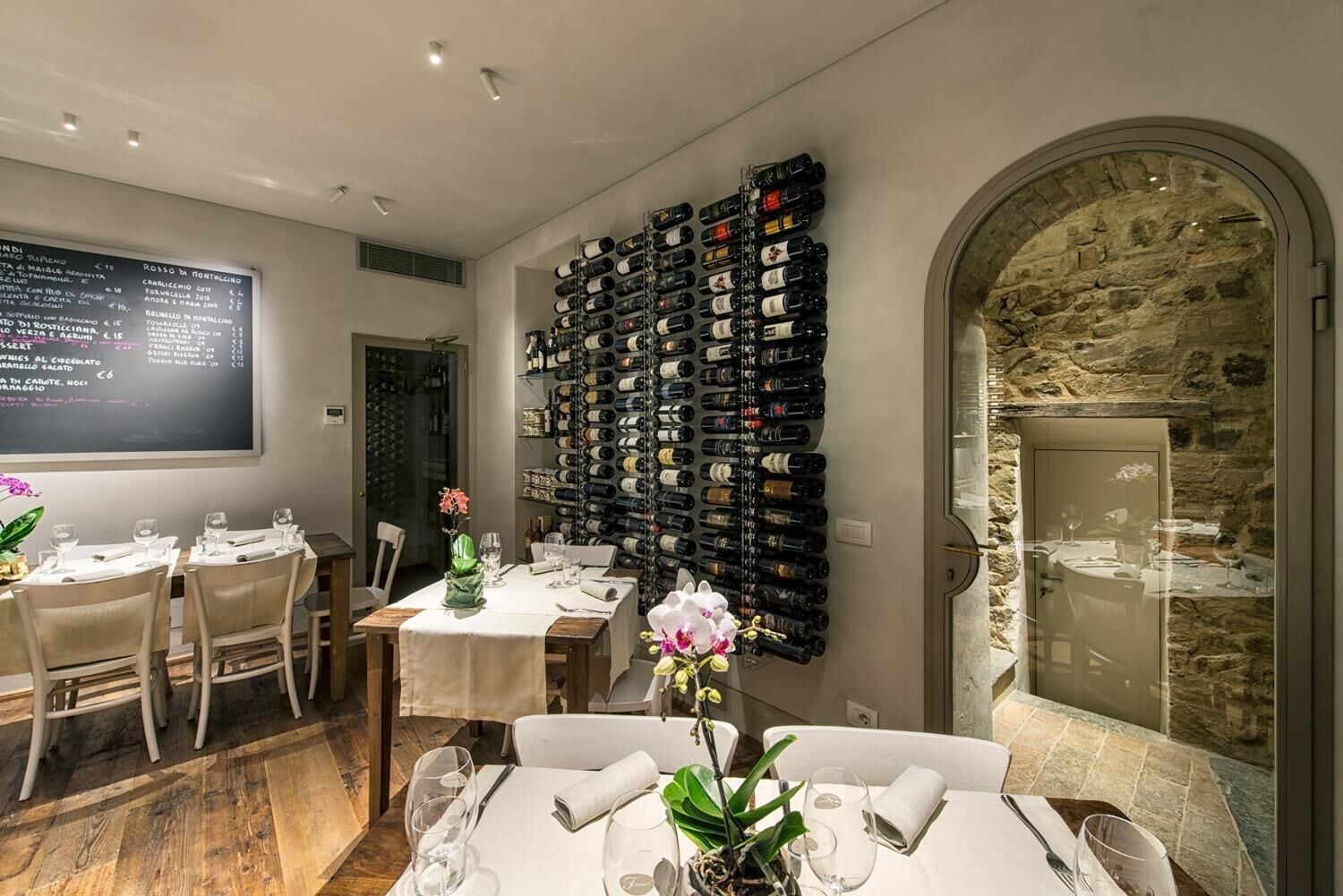 Un interior de restaurante con varias mesas, una pared de vinos y una pared de piedra con puerta de cristal.
