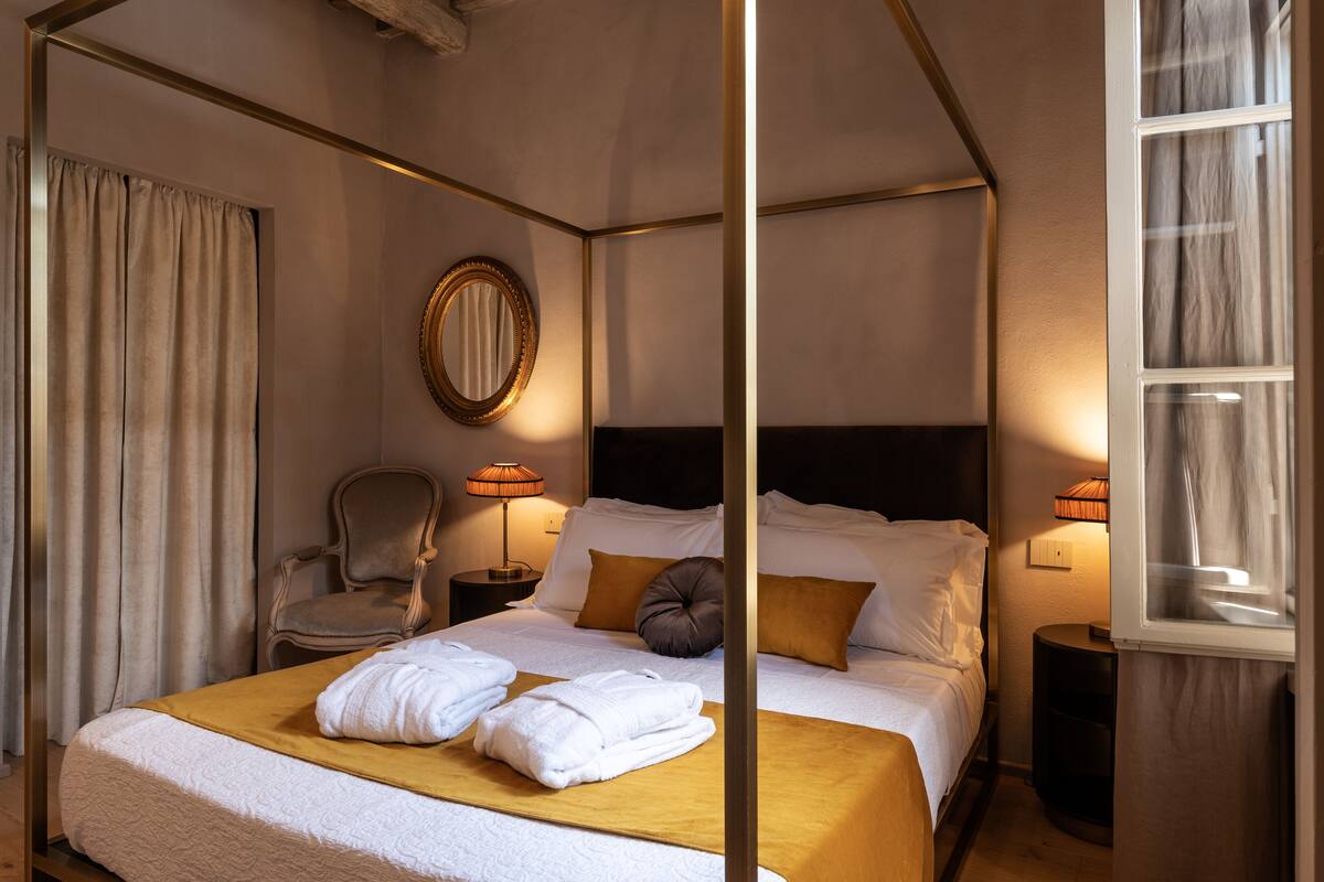 Una habitación de hotel con cama con dosel, un espejo redondo, una silla, dos mesitas de noche con lámparas y un ventanal con cortinas.