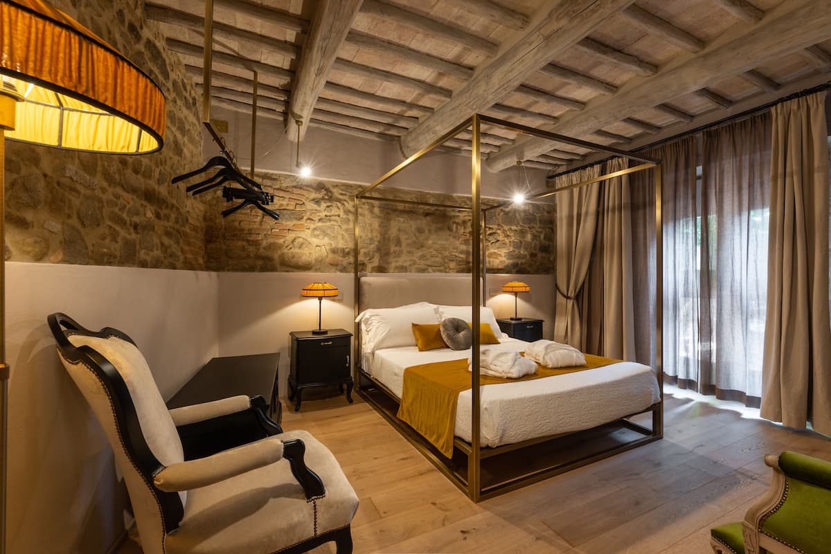 Una cama con dosel con cabecera blanca, un sillón verde, una mesita de noche de madera con una lámpara y una pared de piedra.