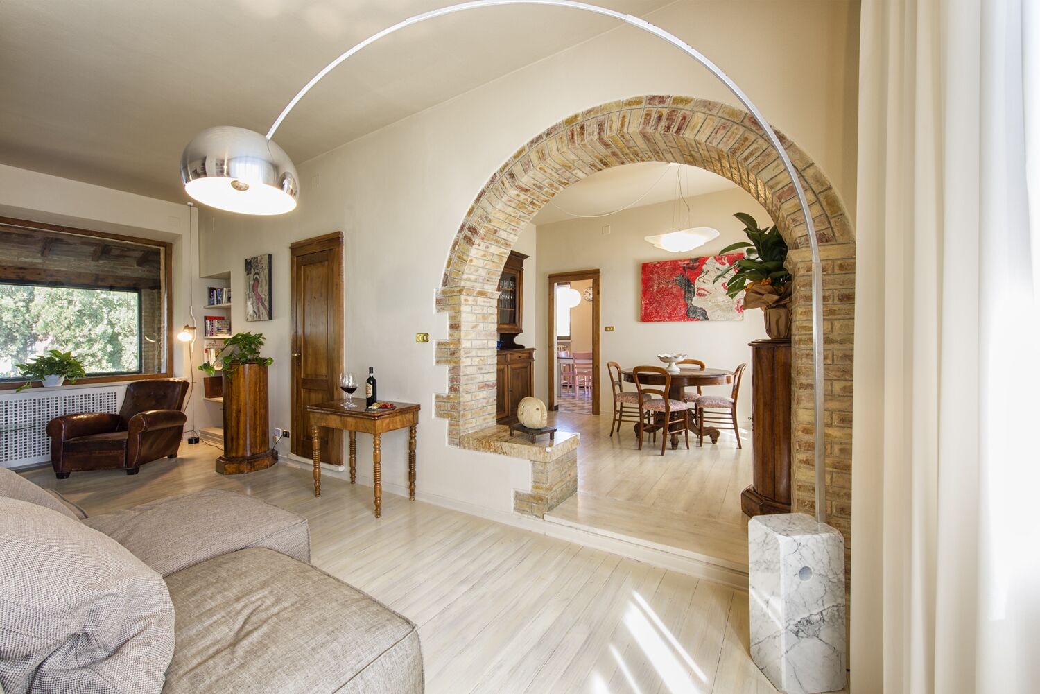 A spacious living room with a large arched doorway leading to a dining area. There is a leather sofa, a wooden table with a wine bottle, and a potted plant.