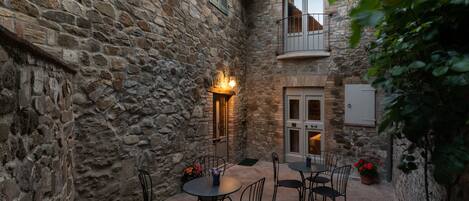 A stone courtyard with a dining area featuring round tables and chairs, a small illuminated wall feature, and a door with a balcony.