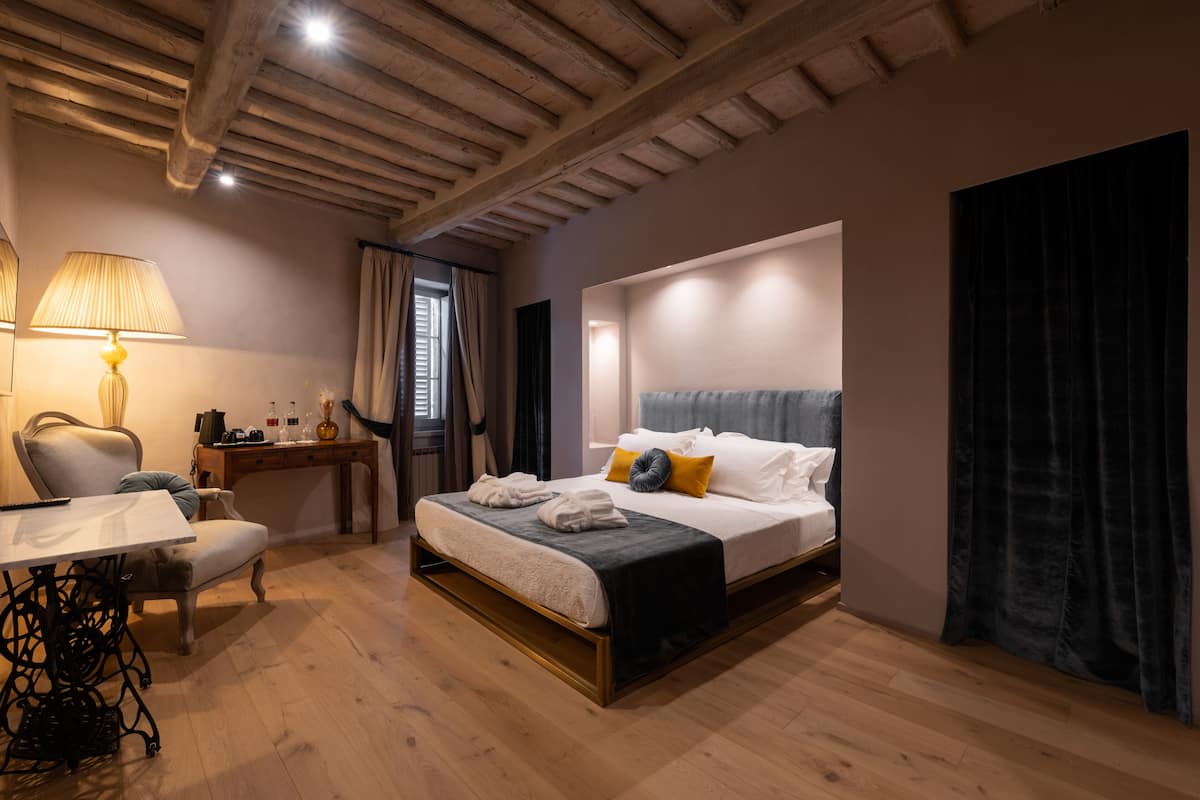 Un dormitorio con techo de madera, cama con ropa de cama blanca y gris, escritorio con lámpara y ventana con cortinas.