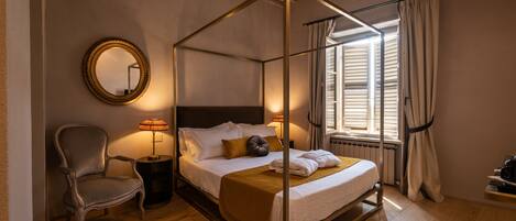 A four-poster bed with a wooden frame, a chair, a round mirror, and a bedside table with a lamp.