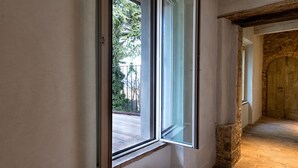 Una escalera de piedra conduce a una habitación con un amplio ventanal, vigas de madera en el techo y un muro de piedra.