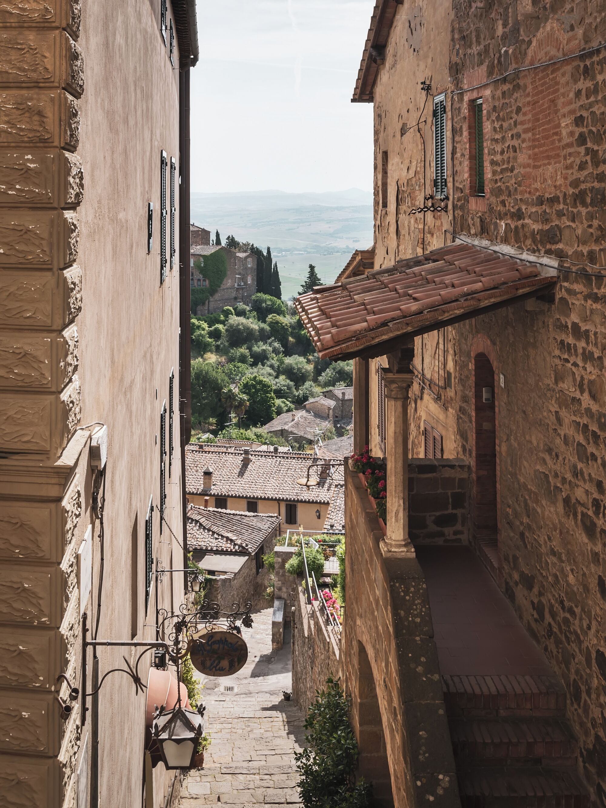 A narrow street with stone buildings, a view of the countryside, and a sign hanging from a balcony.