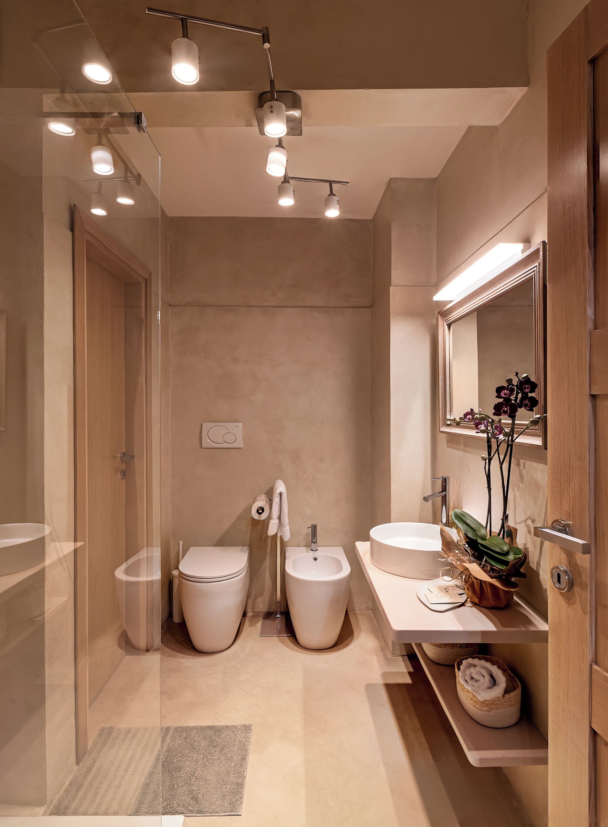 A modern bathroom with a glass-enclosed shower, two toilets, a bidet, a sink, and a mirror with an orchid plant.