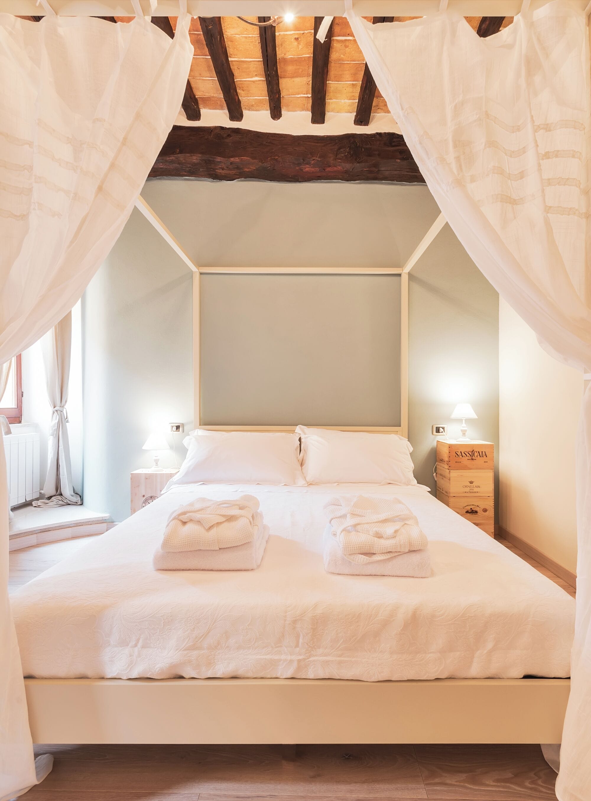 A bedroom with a canopy bed, white linens, two bedside tables with lamps, and a wooden chest of drawers.