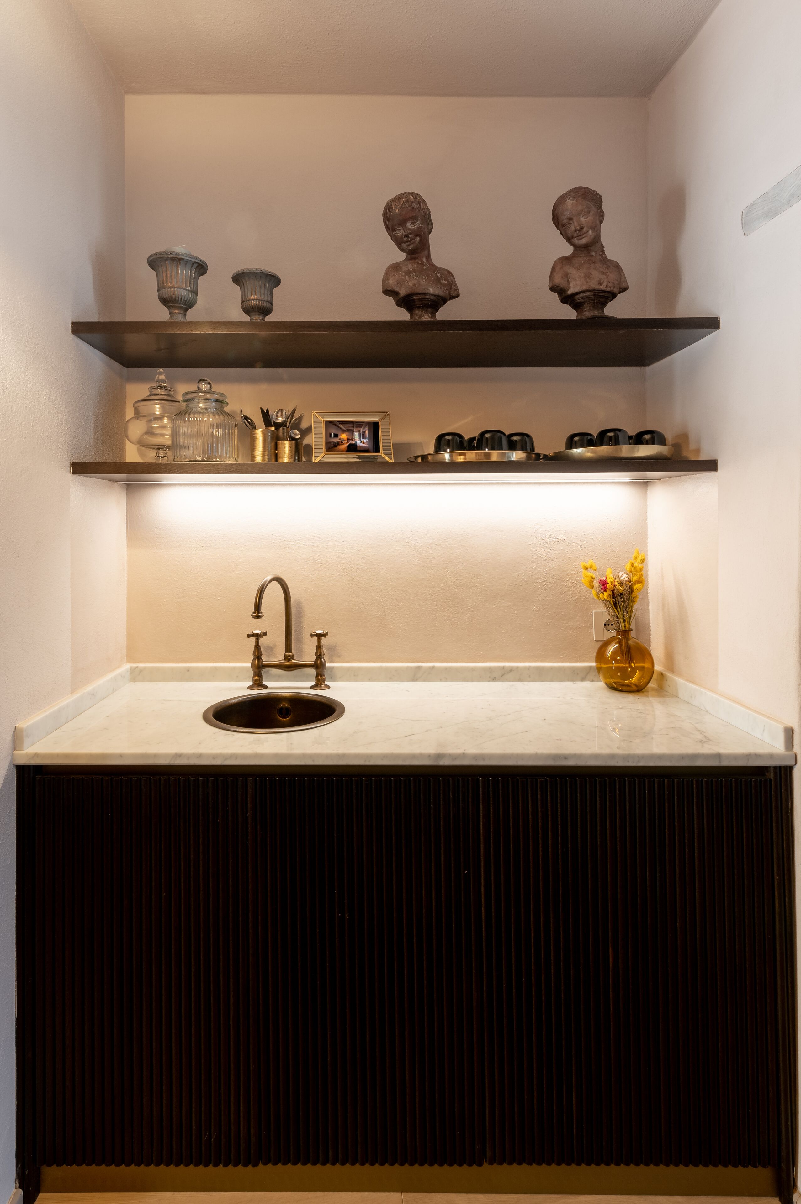 Una cocina con un lavamanos, un jarrón con flores y objetos decorativos en repisas.