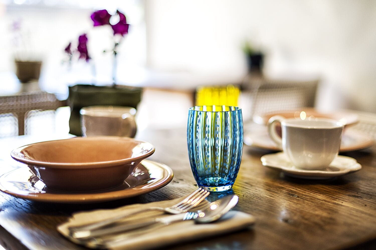 A dining table with a brown plate, a blue glass, a white cup, and a spoon.