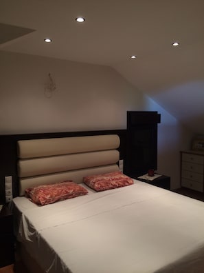A bedroom with a bed, two pillows, a nightstand, a dresser, and recessed ceiling lights.
