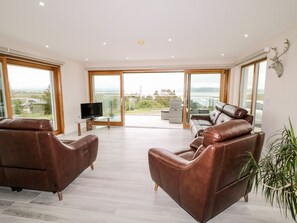 A spacious living room with leather sofas, a flat-screen TV, and a large sliding glass door leading to a balcony with a view of the countryside. - Traeannagh Bay House (Lettermacaward)
