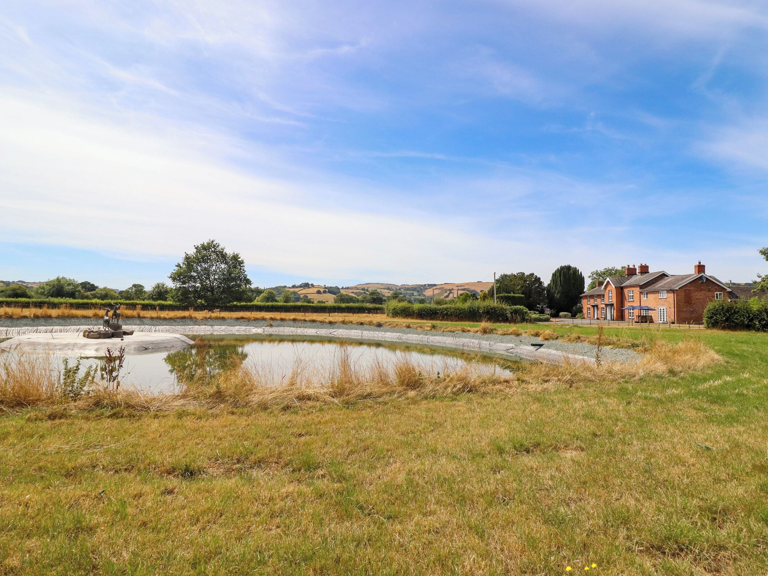 A rural landscape with a pond, a brick house, and rolling hills in the background.