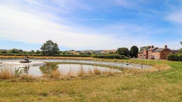 A rural landscape with a pond, a brick house, and rolling hills in the background.