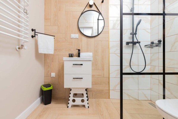A modern bathroom with a white sink, wooden panel walls, a round mirror, a black showerhead, and a white towel on a towel rack.