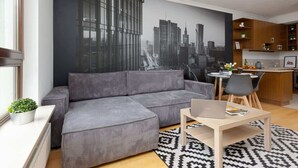 Un salon moderne avec un canapé d’angle gris, un coin repas avec une petite table et des chaises, et une cuisine avec des meubles en bois.