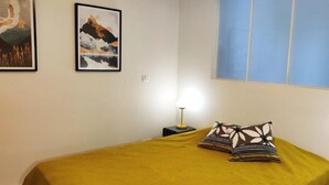 Una habitación con una cama cubierta con un edredón color mostaza, dos almohadas estampadas, una mesita de noche con una lámpara y dos cuadros enmarcados en la pared.