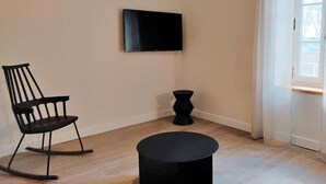 Una habitación con suelo de madera, una mecedora negra, un reposapiés negro, un florero negro y un televisor de pantalla plana montado en la pared.