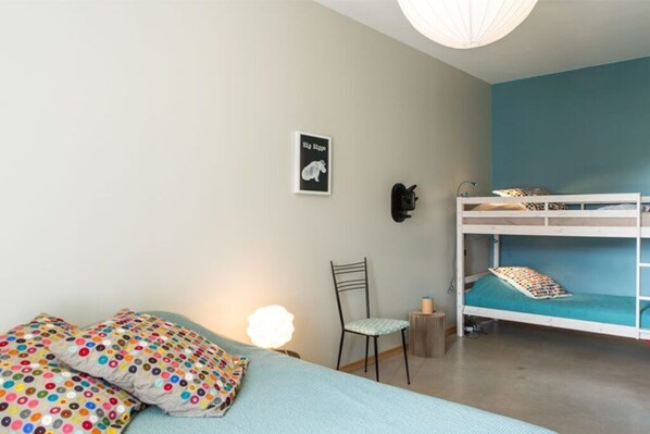 A bedroom with a bunk bed, a chair, a desk, and a wall-mounted lamp.