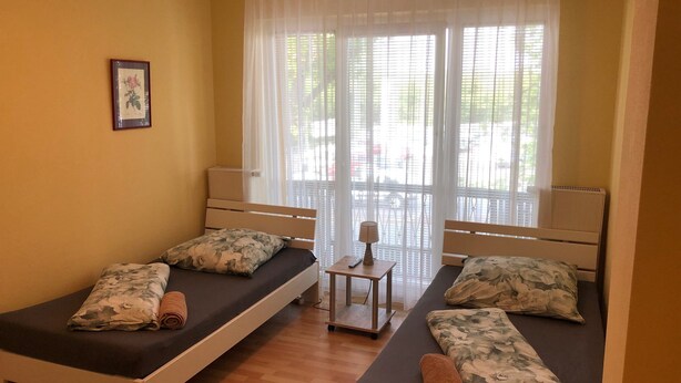 A room with two single beds, a small table with a lamp, and a window with sheer curtains.