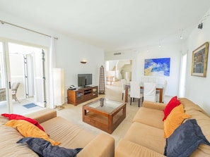 Un salon spacieux comprenant un ensemble de canapés, une table basse et un téléviseur. On y trouve également un coin repas avec des chaises et une table. La pièce est dotée de grandes fenêtres et d’un escalier menant à l’étage supérieur.