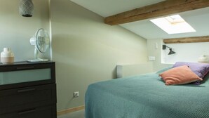 Ein Schlafzimmer mit Dachfenster, einem Bett mit türkisgrünlicher Bettdecke, einer Kommode mit Schubladen, einer Wandleuchte und einer Hängelampe.